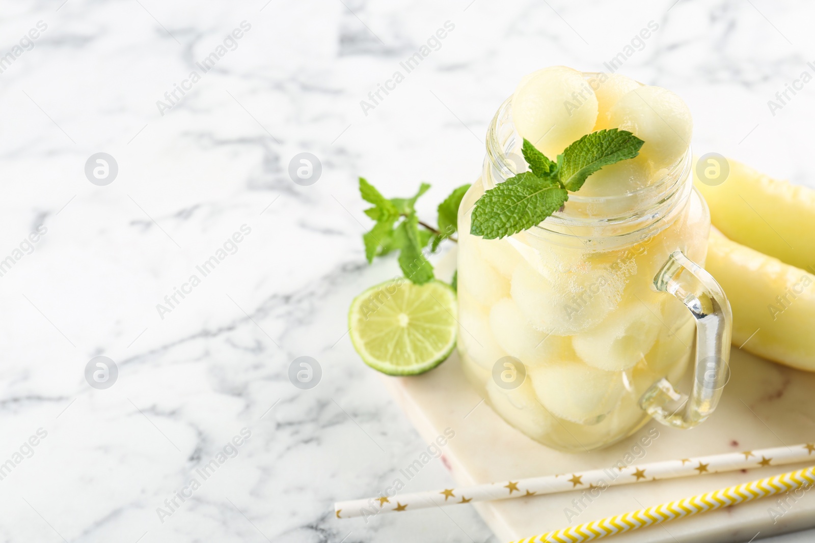 Photo of Mason jar of melon ball cocktail served with mint and lime on white marble table. Space for text