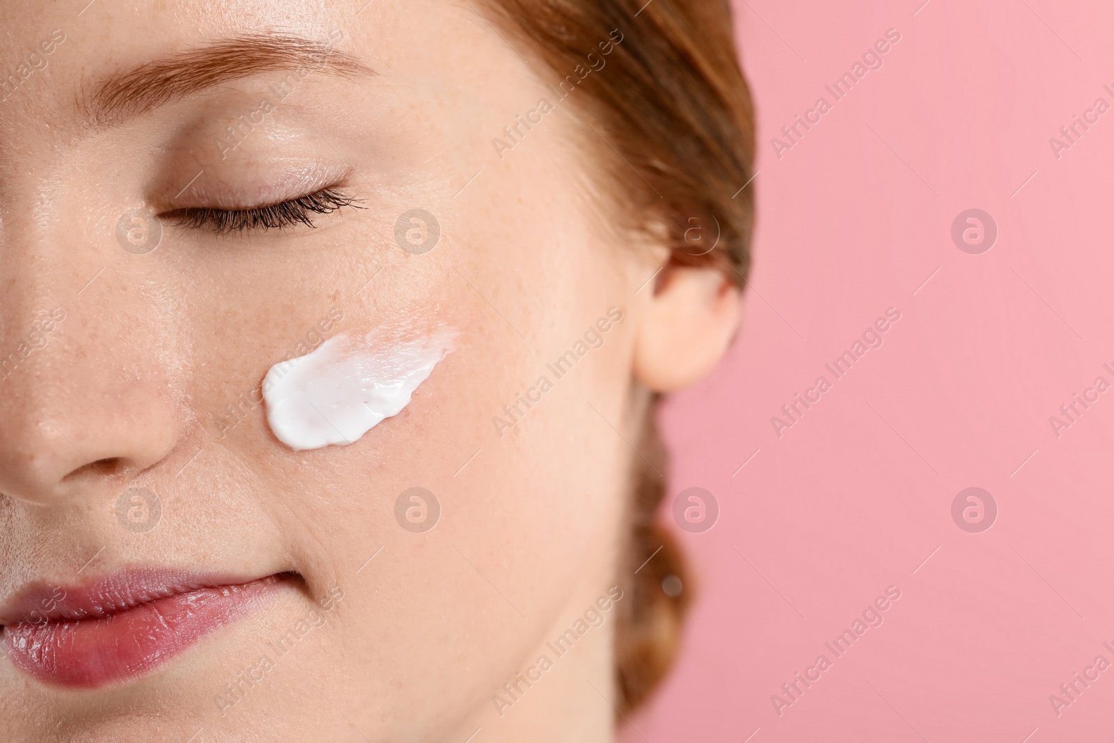 Photo of Beautiful woman with freckles and cream on her face against pink background, closeup. Space for text