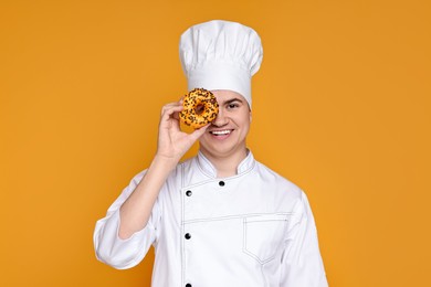 Photo of Portrait of happy confectioner in uniform holding donut on orange background