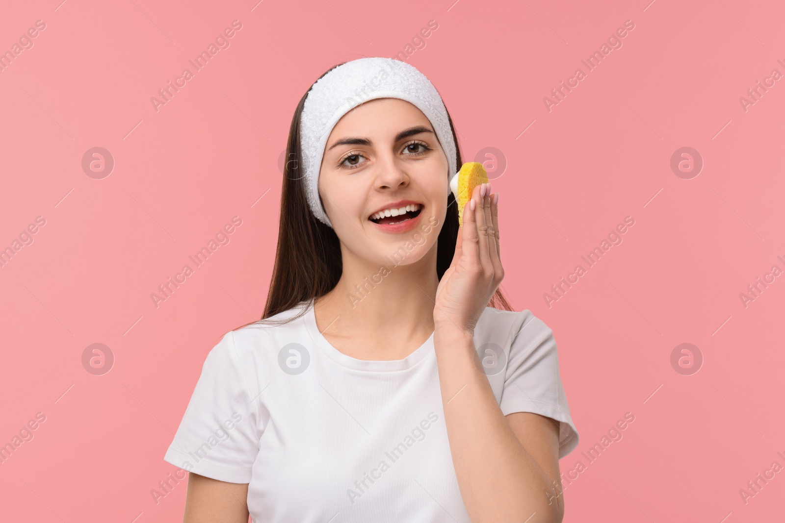 Photo of Young woman with headband washing her face using sponge on pink background