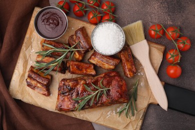 Photo of Tasty roasted pork ribs served with sauce, rosemary and tomatoes on brown table, flat lay