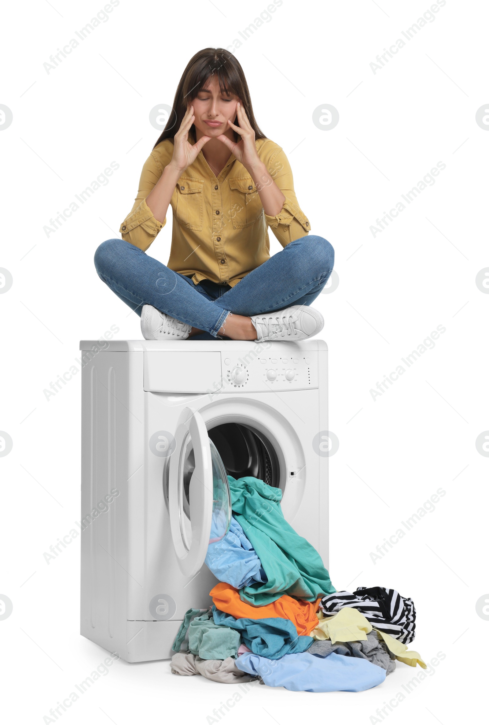 Photo of Confused woman sitting on washing machine with scattered laundry against white background