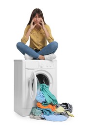 Photo of Confused woman sitting on washing machine with scattered laundry against white background