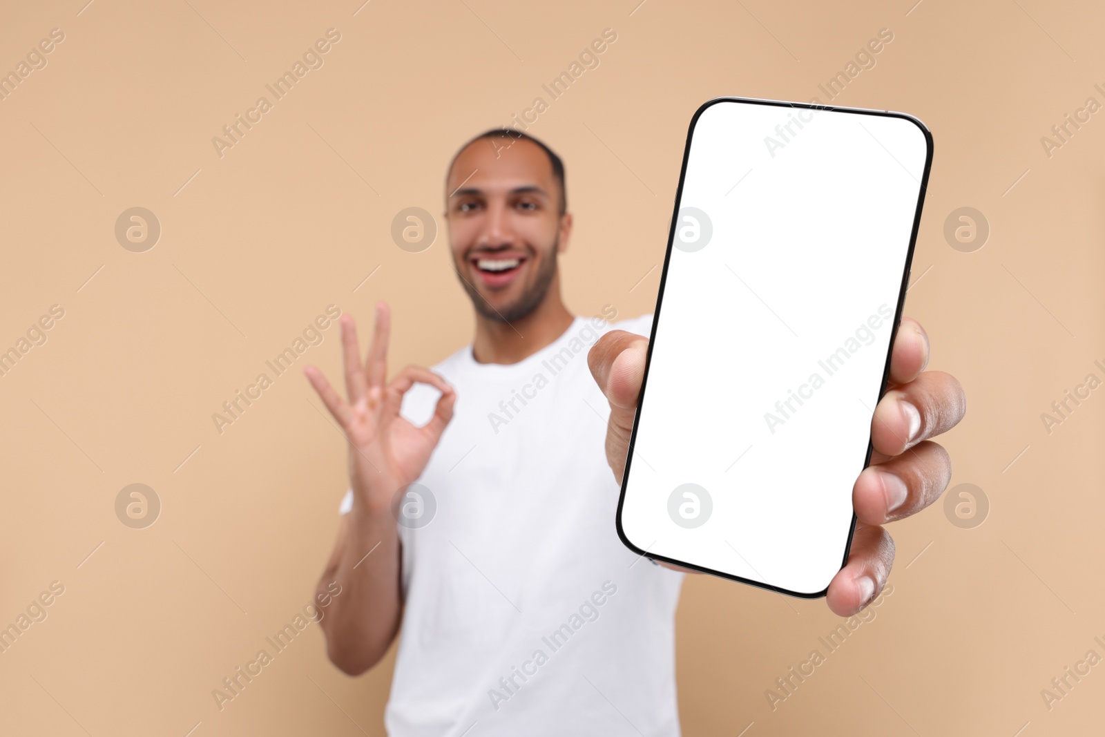 Photo of Young man showing smartphone in hand and OK gesture on beige background, selective focus. Mockup for design