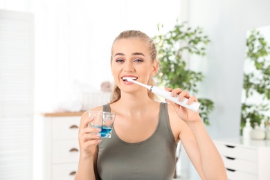 Woman brushing teeth and holding glass with mouthwash in bathroom