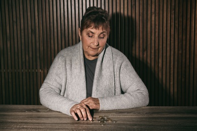 Poor senior woman with coins at table against wooden wall