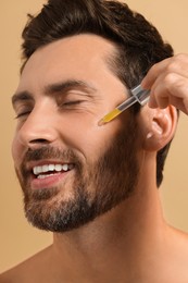 Photo of Smiling man applying cosmetic serum onto his face on beige background, closeup