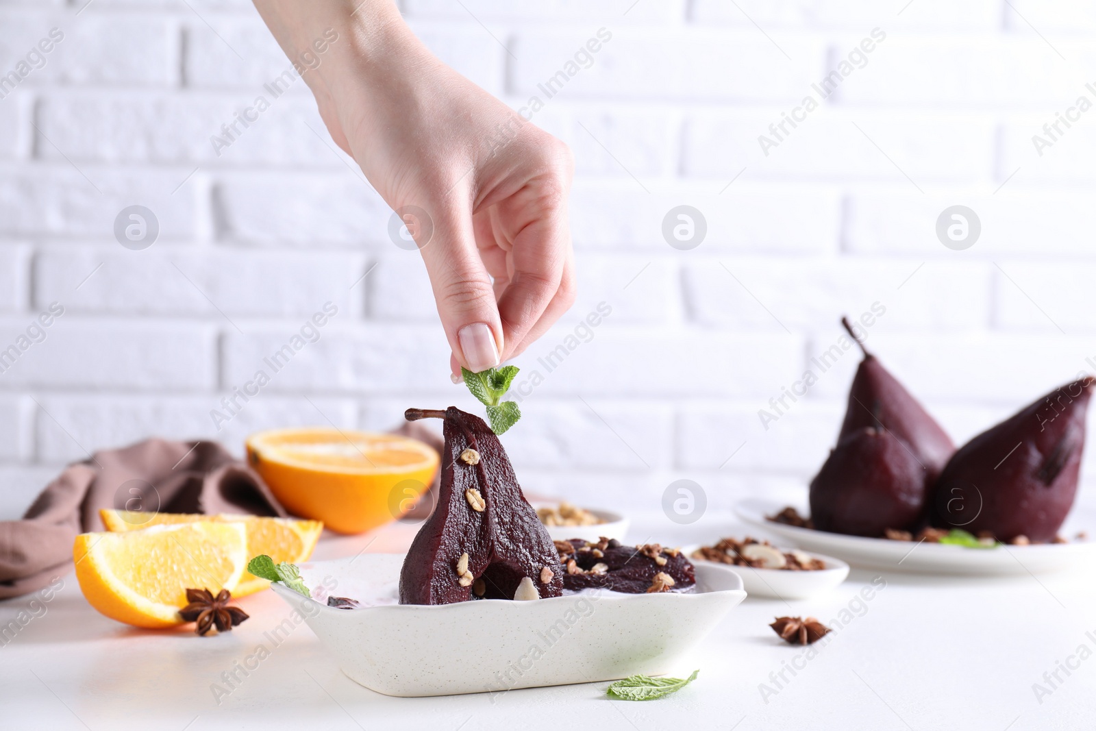 Photo of Woman putting mint on tasty red wine poached pears in bowl at white table, closeup