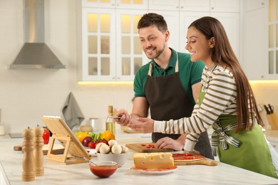 Photo of Happy couple making pizza together while watching online cooking course via tablet in kitchen