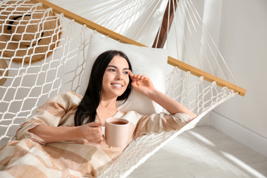 Photo of Young woman with cup of tea in hammock at home