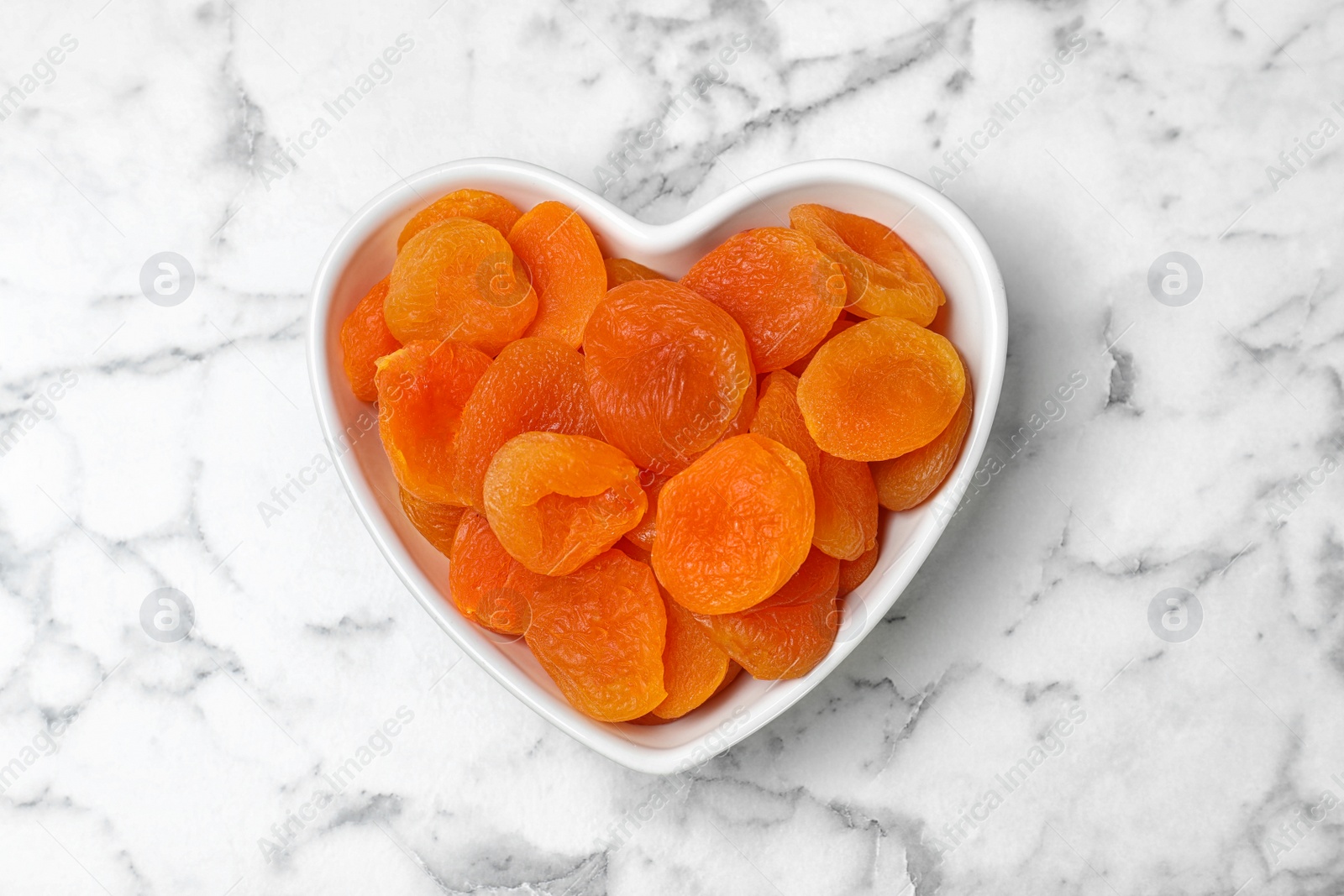 Photo of Heart shaped bowl with apricots on marble background, top view. Dried fruit as healthy food