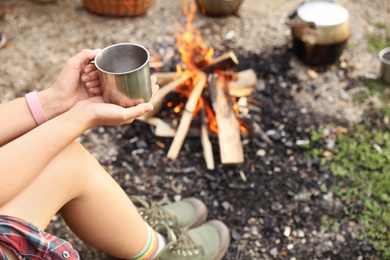 Photo of Young woman with mug near bonfire outdoors, focus on hands. Camping season