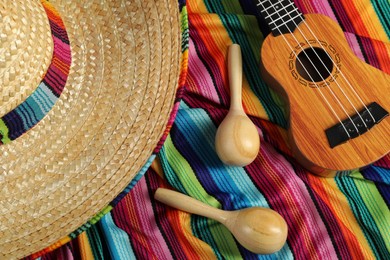 Photo of Mexican sombrero hat, maracas and ukulele on color poncho, top view