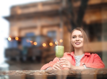 Pretty young woman with cocktail at table in cafe, view from outdoors through window