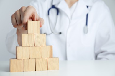 Photo of Doctor building pyramid of blank wooden cubes on white table against light background, closeup. Space for text