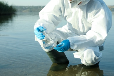 Scientist in chemical protective suit with conical flask taking sample from river for analysis, closeup