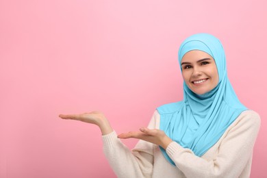 Portrait of Muslim woman in hijab pointing at something on pink background, space for text