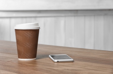 Photo of Cardboard coffee cup with lid and phone on wooden table. Space for text