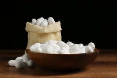 Photo of White silk cocoons with bowl and sackcloth bag on wooden table, closeup