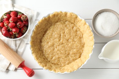 Photo of Making shortcrust pastry. Raw dough in baking dish, strawberries, sugar, rolling pin and milk on white wooden table, top view