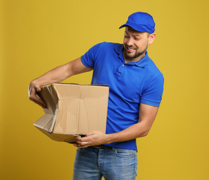 Photo of Emotional courier with damaged cardboard box on yellow background. Poor quality delivery service