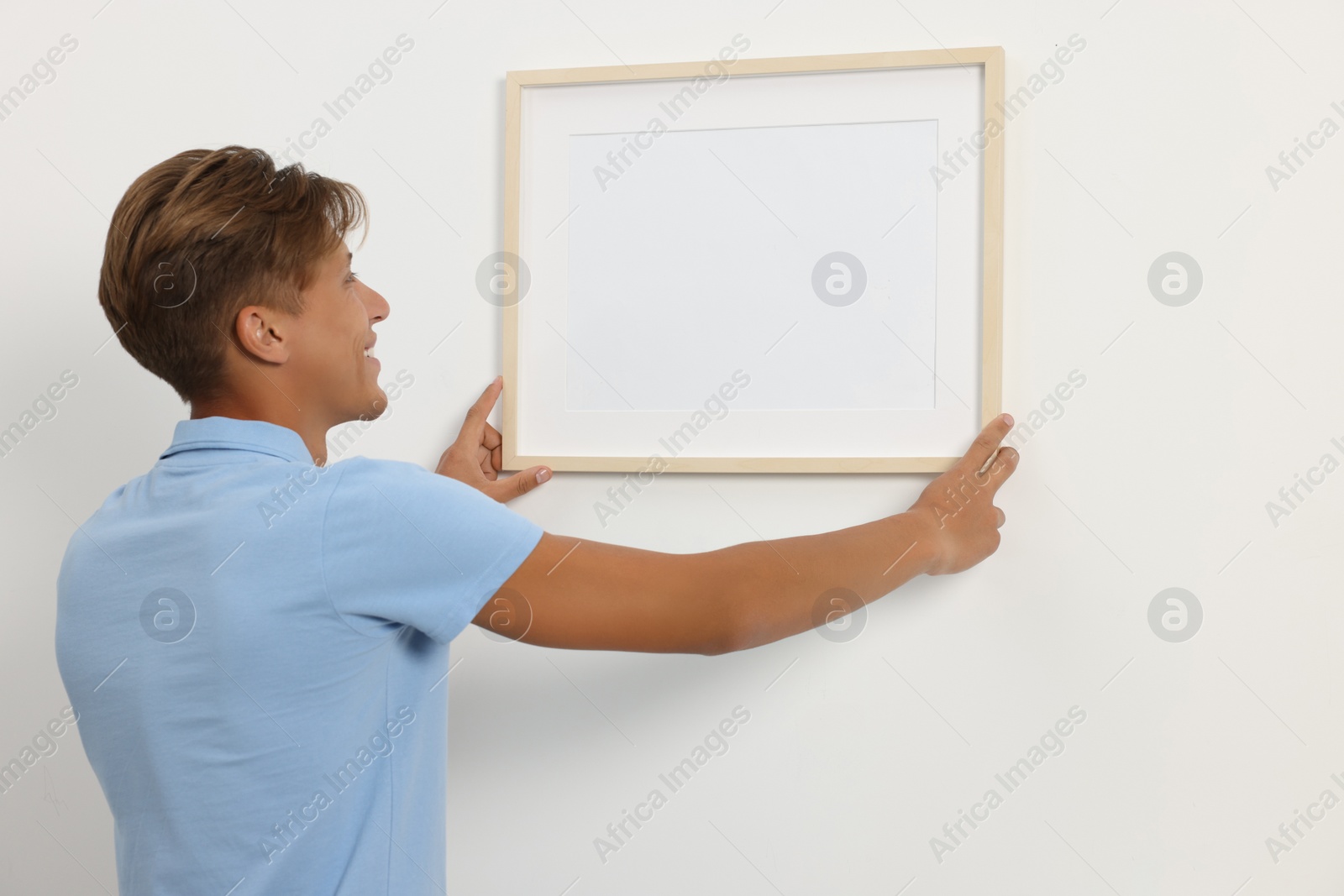 Photo of Young man hanging picture frame on white wall indoors