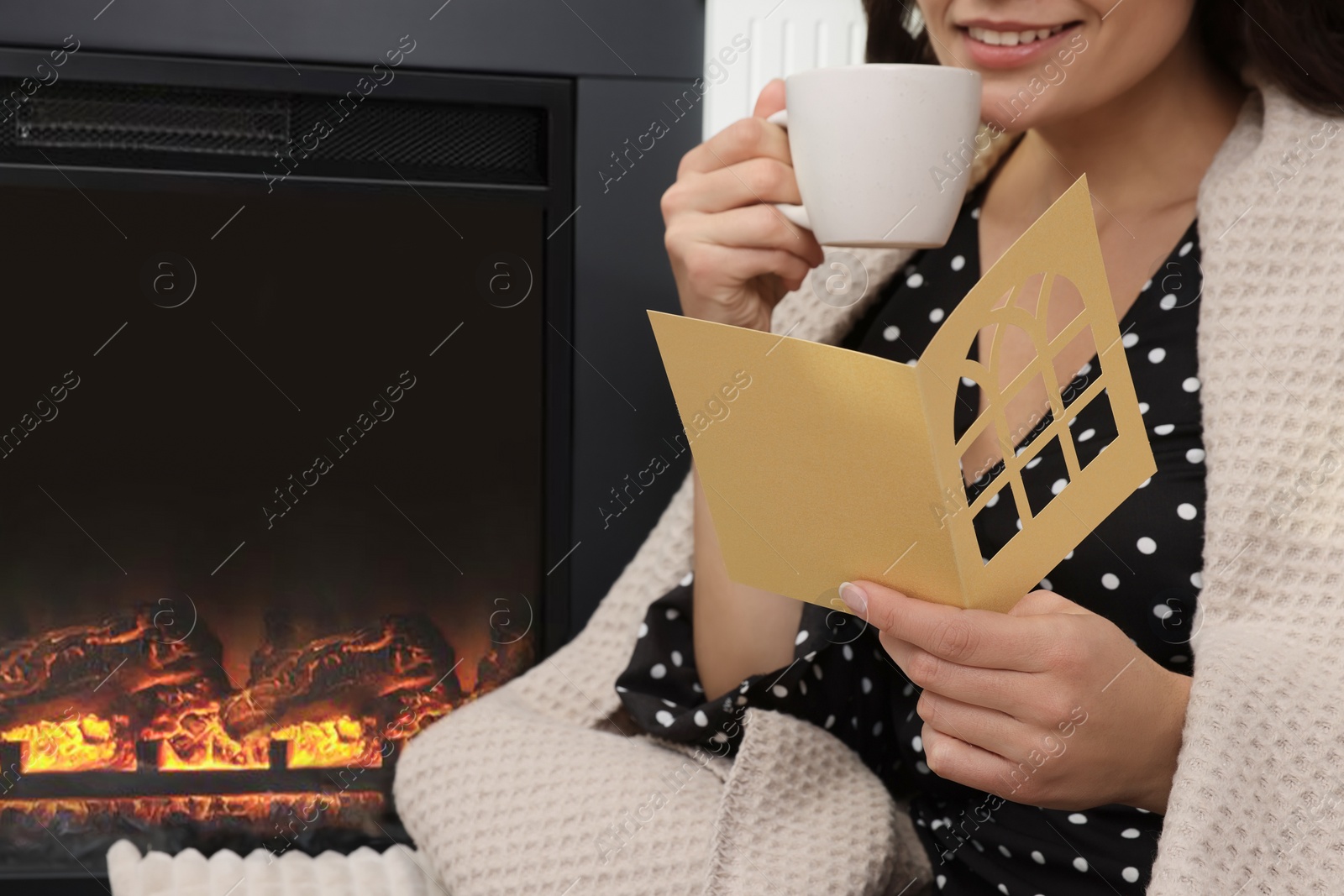 Photo of Young woman sitting near fireplace with greeting card and hot drink indoors, closeup. Space for text