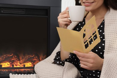 Photo of Young woman sitting near fireplace with greeting card and hot drink indoors, closeup. Space for text