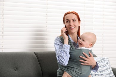 Mother talking on smartphone while holding her child in sling (baby carrier) indoors