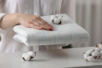 Photo of Woman taking bed sheet and cotton flowers on white wooden table in room, closeup