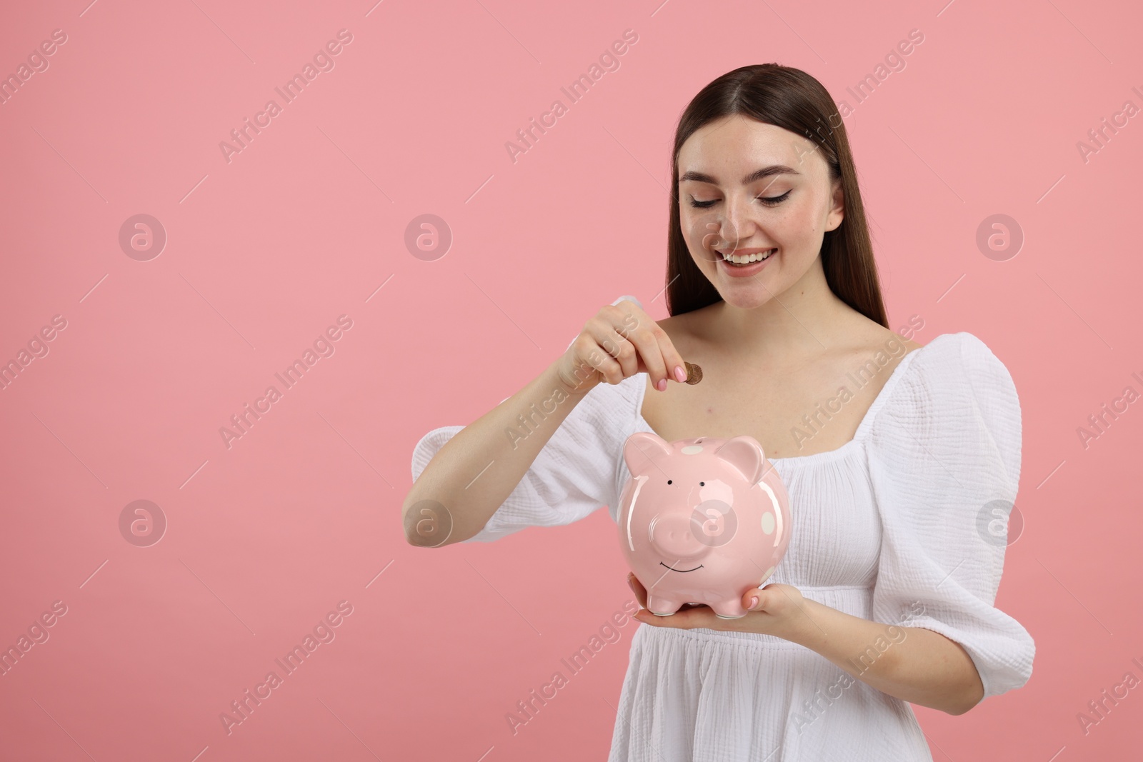 Photo of Happy woman putting coin into piggy bank on pink background, space for text