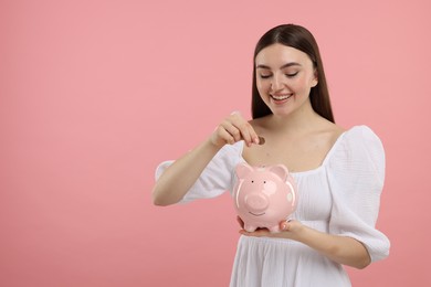 Happy woman putting coin into piggy bank on pink background, space for text