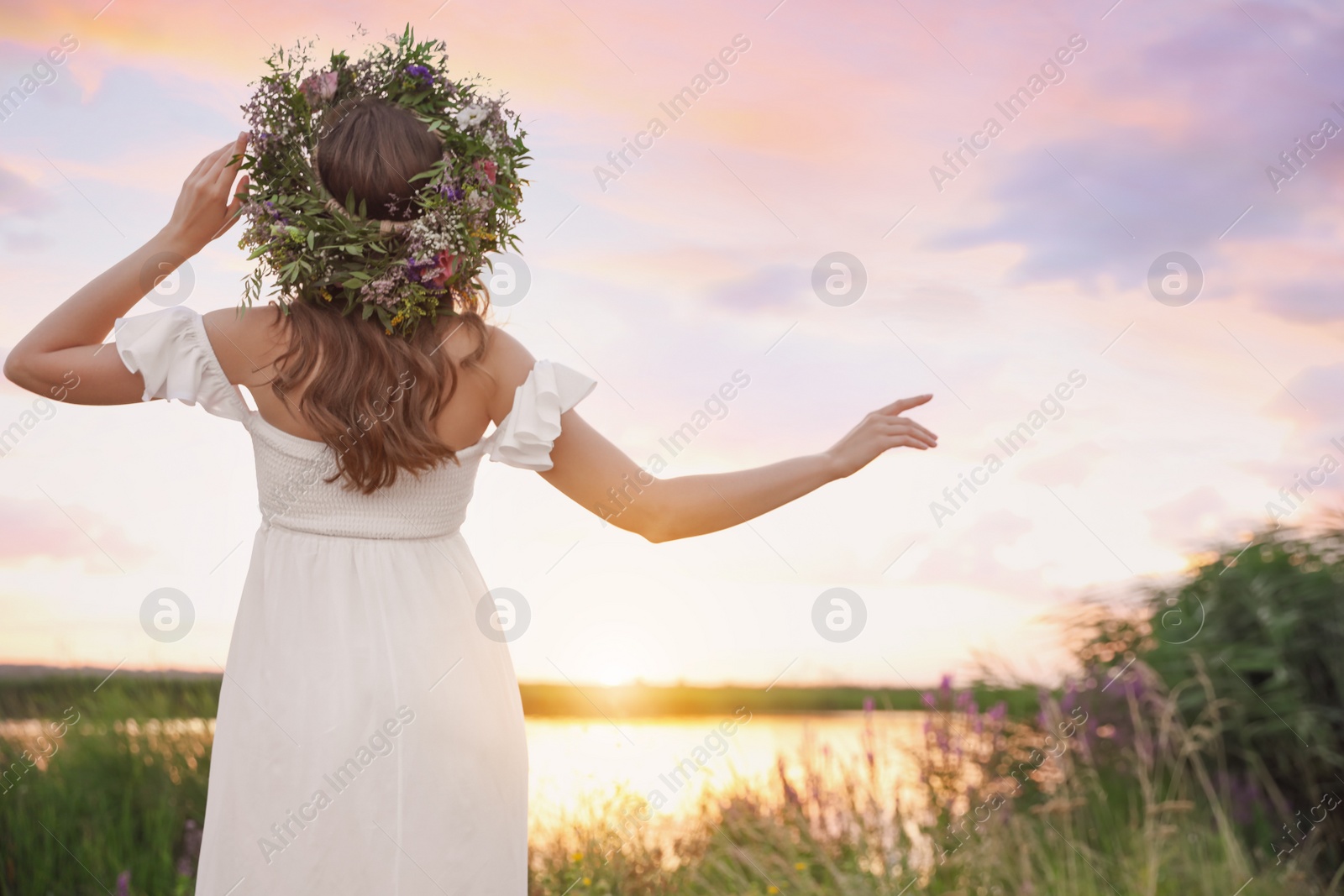 Photo of Young woman wearing wreath made of beautiful flowers outdoors at sunset, back view