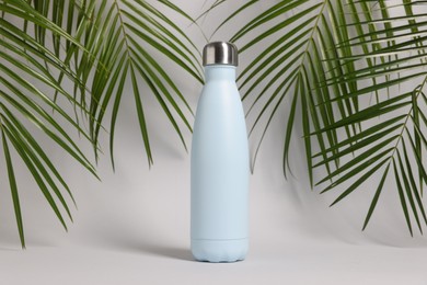 Stylish thermo bottle near tropical leaves on light grey background