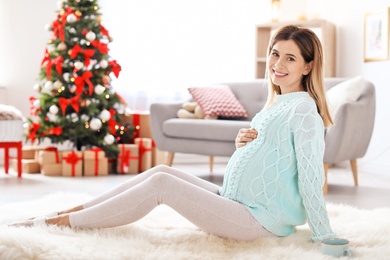 Photo of Happy pregnant woman sitting on floor in room decorated for Christmas