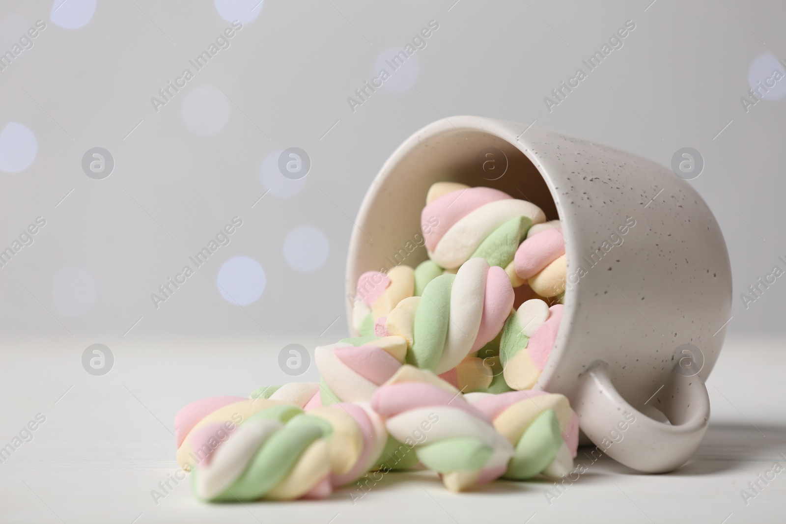 Photo of Ceramic cup with colorful marshmallows on white table against blurred lights, closeup