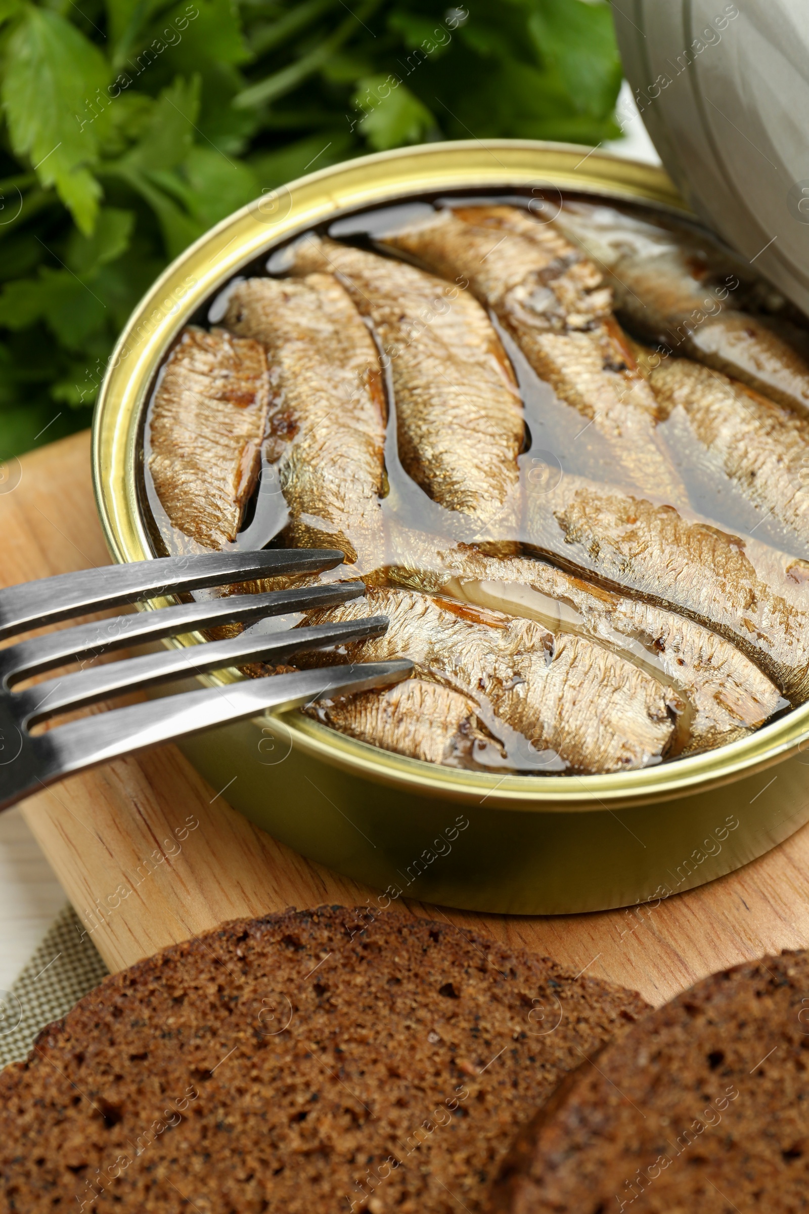 Photo of Fork with canned sprats and bread on wooden board, closeup