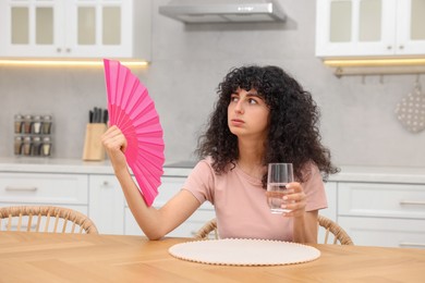 Photo of Young woman with glass of water waving pink hand fan to cool herself at table in kitchen
