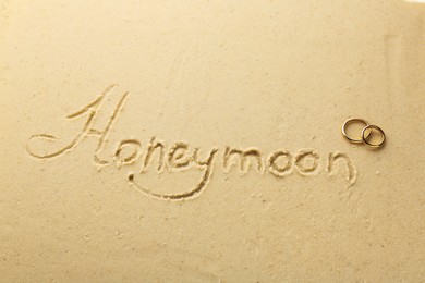Photo of Word Honeymoon written on sand and two golden rings, above view