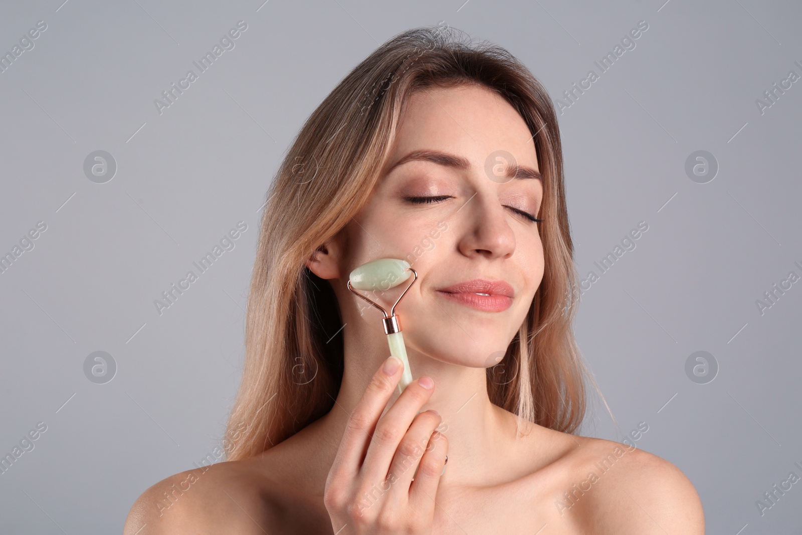 Photo of Young woman using natural jade face roller on light grey background