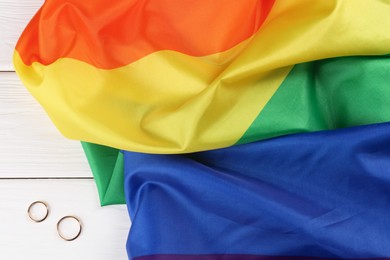 Rainbow LGBT flag and wedding rings on white wooden table, top view