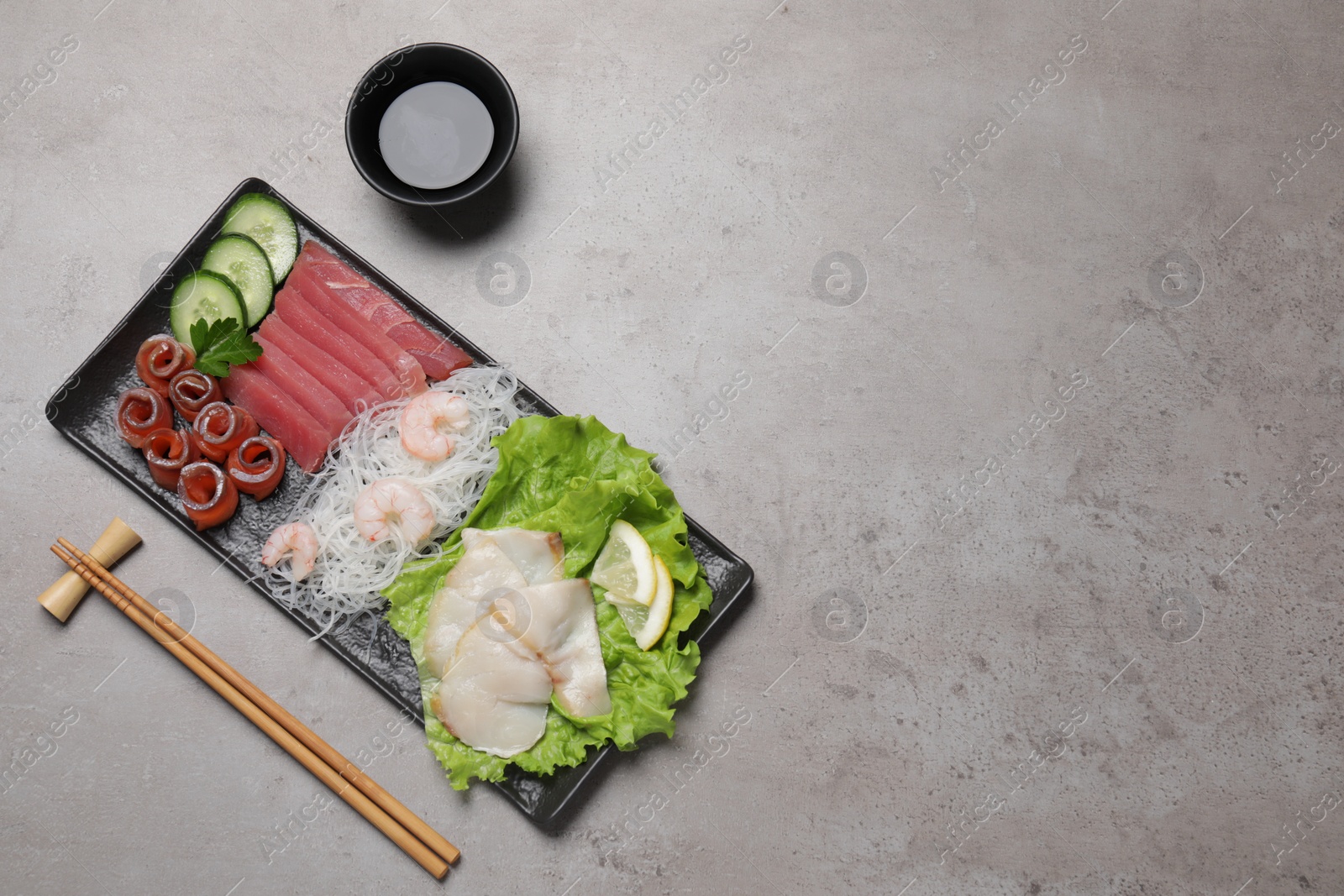 Photo of Sashimi set (raw slices of tuna, oily fish, salmon and shrimps) served with cucumber, lettuce, funchosa, soy sauce and lemon on light grey table, flat lay