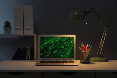 Modern laptop near lamp and holder with stationery on table at night