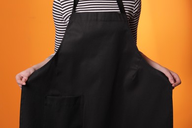 Photo of Woman putting on black apron against orange background, closeup