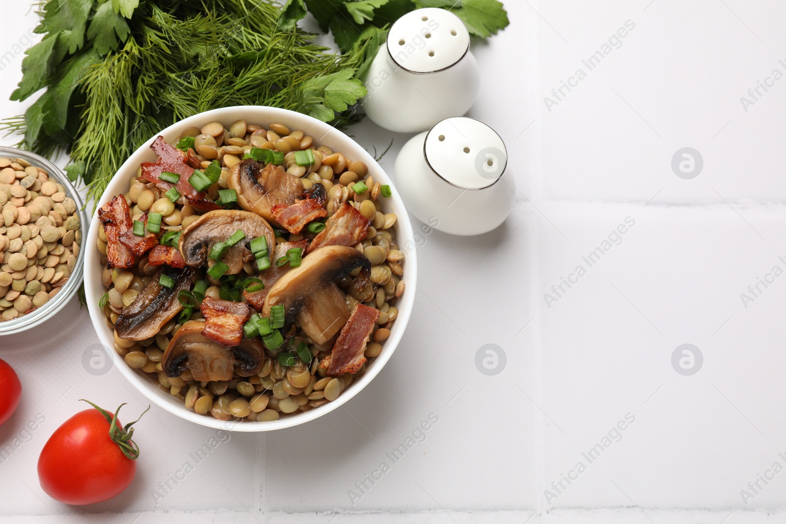 Photo of Delicious lentils with mushrooms, bacon and green onion in bowl served on white tiled table, flat lay. Space for text