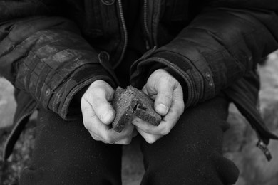 Photo of Poor homeless man holding piece of bread outdoors, closeup. Black and white effect