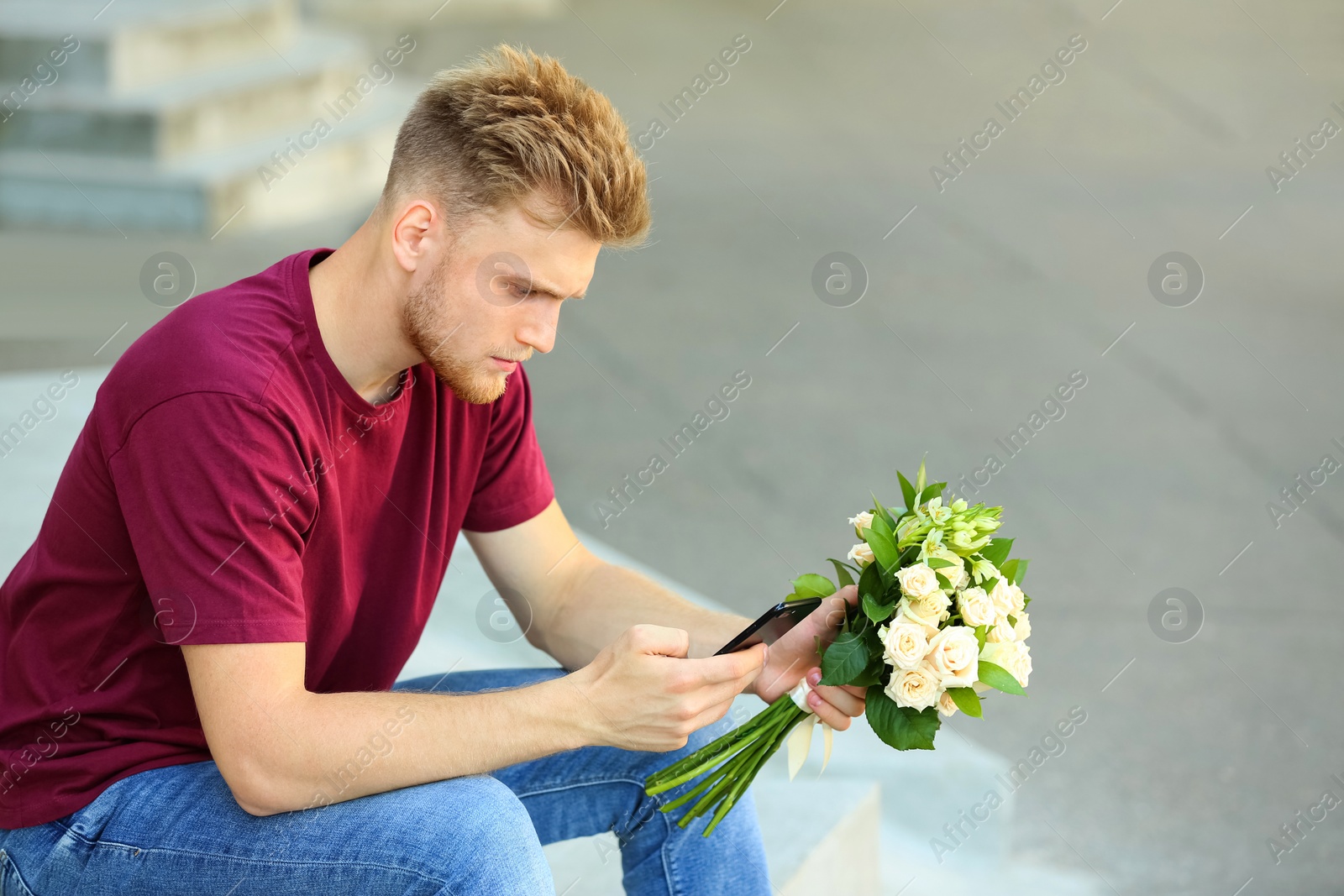 Image of Young man with bouquet of flowers and phone waiting for his girlfriend outdoors