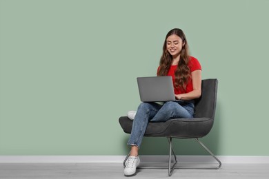 Happy woman with laptop sitting in armchair near pale green wall indoors. Space for text