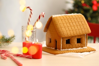 Photo of Tasty gingerbread house and burning candles on light table indoors