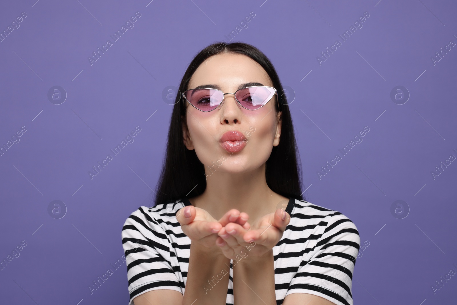 Photo of Beautiful young woman in stylish sunglasses blowing kiss on purple background